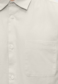 Gros plan sur le devant d’une chemise beige clair boutonnée, avec col, quatre boutons visibles et une poche poitrine sur le côté droit.