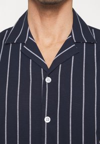 Homme portant une chemise bleu marine avec des rayures verticales blanches et deux boutons blancs visibles, montrant l'encolure et une partie du col.
