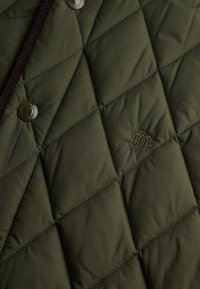 Veste matelassée verte avec un motif en losange, arborant un logo et des boutons-pression en métal, ainsi qu'un cordon en cuir marron contrastant.