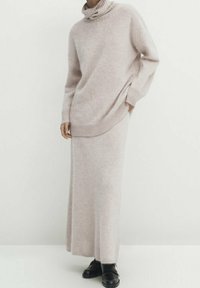 Ljusrosa stickad tröja med hög, räfflad krage, i kombination med en matchande lång kjol. Båda har en mjuk, texturerad yta. Svarta skor bärs.