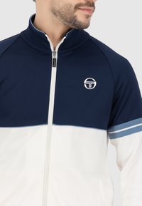 Veste zippée bleu marine et blanche avec un col haut, présentant un logo sur la poitrine gauche et des rayures bleues sur les manches. Texture de tissu lisse.