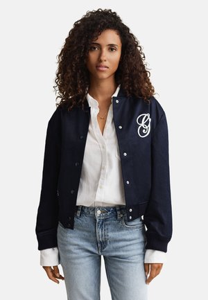 Donna con capelli ricci che indossa una giacca bomber blu navy con emblema bianco, camicia bianca abbottonata e jeans chiari, in piedi davanti a uno sfondo uniforme.