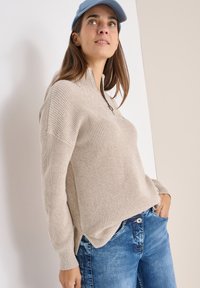 Beżowy sweter dzianinowy z wysokim kołnierzem i zamkiem, w połączeniu z niebieskimi dżinsami. Model nosi jasnoniebieską czapkę, prezentując luźny styl.