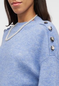 Γυναίκα που φοράει ένα ανοιχτό μπλε πουλόβερ με τέσσερα γυαλιστερά κουμπιά στον ώμο και ένα μονό σειρό λευκό μαργαριταρένιο κολιέ.