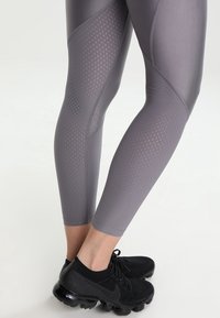 Szare legginsy sportowe z siateczkowymi wstawkami, charakteryzujące się dopasowanym krojem i geometrycznymi wzorami. W połączeniu z czarnymi butami sportowymi.