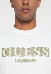 Suumi inimene, kes kannab valget särki, millel on tekstureeritud beež tekst "GUESS" ja "EST 1981" rinnal.