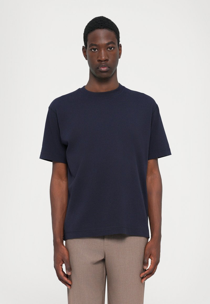 T-shirt en coton bleu marine à manches courtes, col rond et coupe décontractée. Texture lisse, design minimal, associé à un pantalon beige.