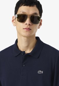 Polo marine en tissu texturé, avec trois boutons et logo crocodile Lacoste. Le modèle porte des lunettes de soleil oversize avec une monture claire et marron.