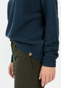 Marineblauwe geribde trui met een relaxte pasvorm en textuur gebreid. Het heeft een klein leren label aan de zoom. Gecombineerd met olijfgroene broek.