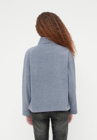 Ανοιχτό μπλε φλις πουλόβερ με ψηλό γιακά, χαλαρούς ώμους και μακριά μανίκια. Ομαλή υφή με άνετη εφαρμογή, συνδυασμένο με σκούρο τζιν.