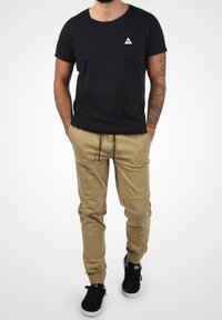 Man som bär en svart t-shirt, beige cargobyxor med snörning, svarta sneakers och en tatuerad ärm på sin högra arm, med händerna i fickorna.