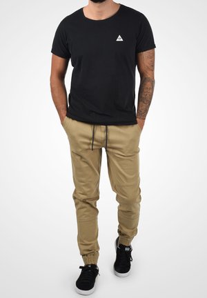 Man wearing a black t-shirt, beige cargo pants with drawstring, black sneakers, and a tattoo sleeve on his right arm, hands in pockets.