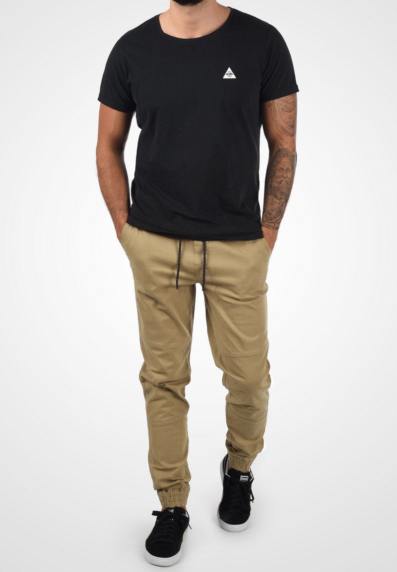 Man som bär en svart t-shirt, beige cargobyxor med snörning, svarta sneakers och en tatuerad ärm på sin högra arm, med händerna i fickorna.