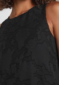 Svart ärmlös blus med en texturerad blommönster, subtil lyster och hög halsringning. Tyget verkar mjukt och lätt.