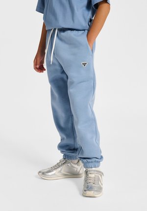 Lyseblå joggingbukser med elastiske manchetter, snøre i taljen og sidelommer; har en lille sort logo på venstre lår. Sølvfarvede sneakers.