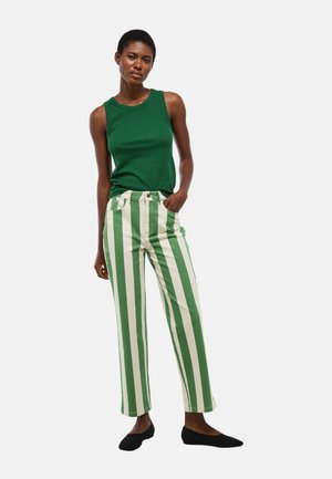 Femme portant un haut vert sans manches, un pantalon à rayures verticales vertes et blanches taille haute, et des chaussures plates noires, debout avec une main dans la poche.