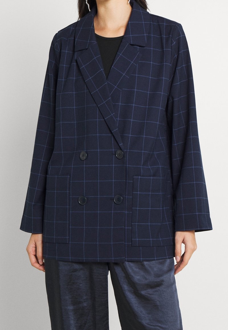 Femme portant un blazer croisé bleu marine avec un motif à carreaux subtil, associé à un pantalon sombre, brillant et texturé.