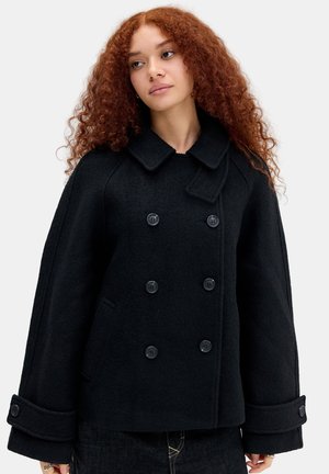 Femme aux cheveux roux bouclés portant un manteau noir croisé à larges manches et poignets boutonnés sur un fond uni.
