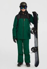 Person in green and black ski outfit, wearing helmet and goggles, holding a snowboard upright with bindings visible.