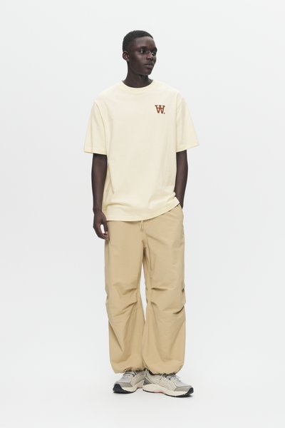 Giovane uomo in piedi, con pantaloni oversize beige, t-shirt oversize crema con un piccolo logo marrone e sneakers beige su sfondo bianco.