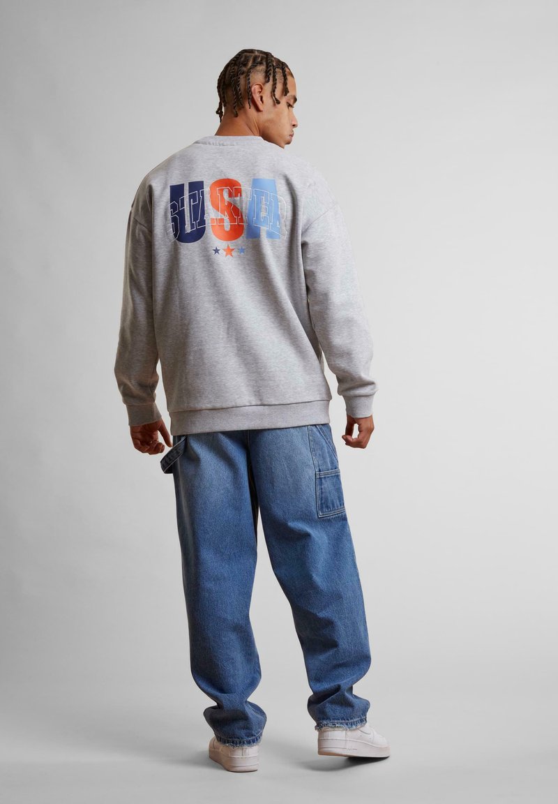 Felpa grigia con grafica "BUSTER" in arancione, blu e blu scuro sul retro. Indossata con pantaloni in denim a vestibilità ampia e scarpe bianche.