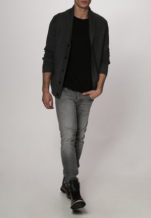 Mies kävelee eteenpäin yllään tummanharmaa napitettu neuletakki, musta paita, vaaleanharmaat farkut ja mustat nahkasaappaat yksinkertaisella taustalla.