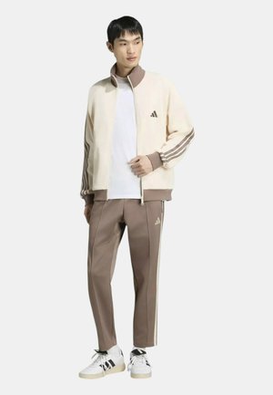 Uomo che indossa una giacca da tuta Adidas color crema e marrone e pantaloni da tuta marroni abbinati con scarpe da ginnastica bianche e nere, in piedi e guarda verso destra.