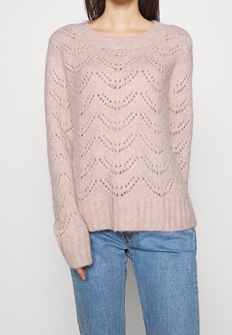 Femme portant un pull en maille rose pâle avec un motif œillet en chevron et un jean denim bleu clair, debout devant un fond uni.