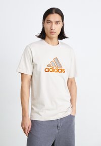 Tricou din bumbac crem, cu logo Adidas brodat portocaliu, guler rotund și mâneci scurte, asortat cu pantaloni scurți gri.