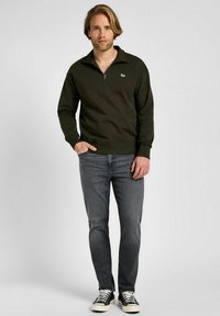 Felpa zip-up verde scuro con colletto, abbinata a jeans grigi e sneaker neri. Presenta un piccolo logo sul petto. Tessuto morbido.