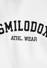 Tissu blanc avec un texte noir en gras qui indique "SMILODON" en arc de cercle avec "ATHL. WEAR" en dessous dans une police plus petite.