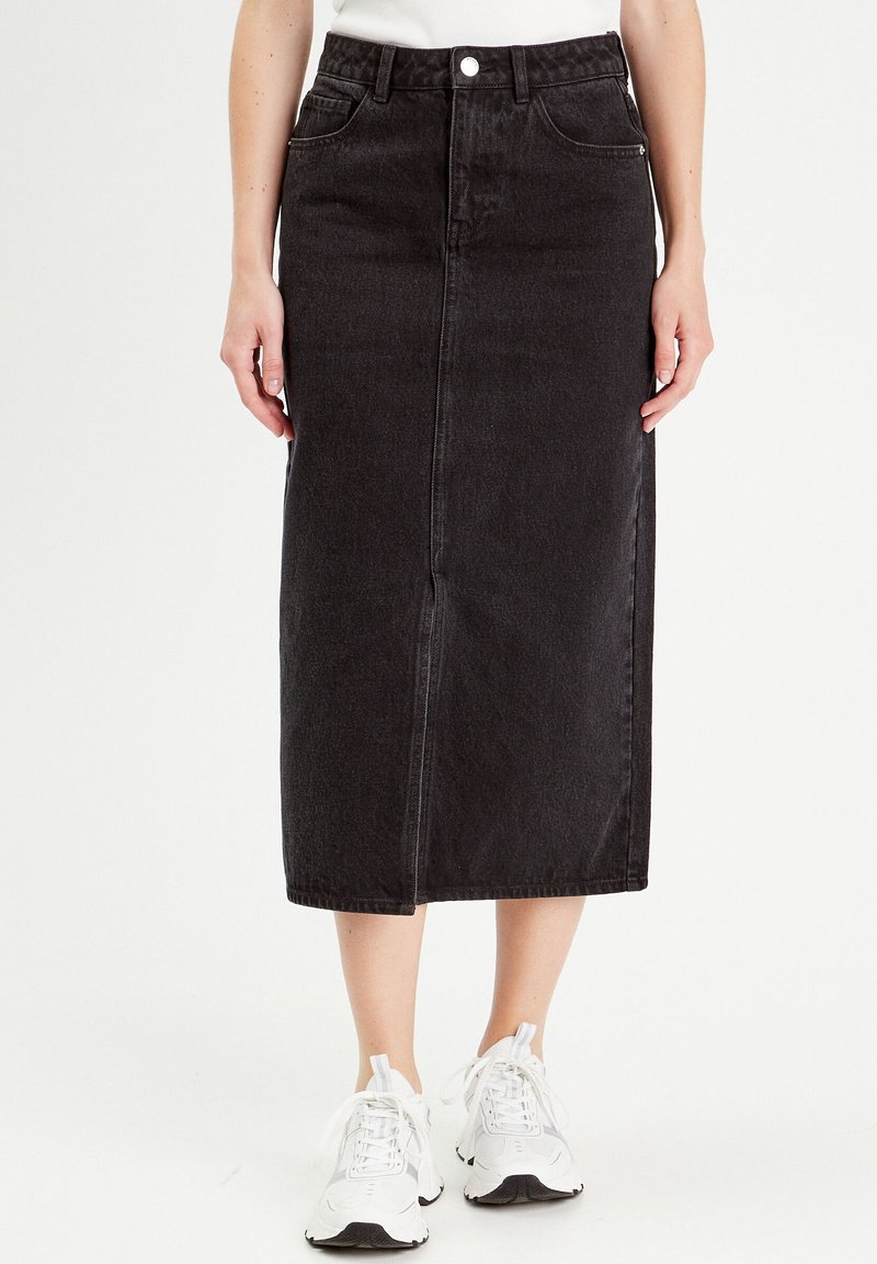 Jupe midi en denim noir avec une fente frontale, cinq poches et une fermeture à bouton. Associée à des baskets blanches pour un look décontracté.