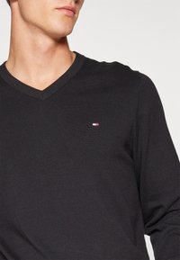 Maglietta nera a maniche lunghe con scollo a V, realizzata in tessuto a costine, con un piccolo logo in rosso, bianco e blu sul lato sinistro del petto.