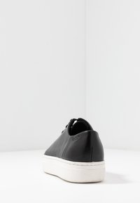 Zapatilla de cuero negra con parte trasera redondeada, suela de goma blanca y un diseño minimalista. Presenta cordones y una sutil marca en el talón.