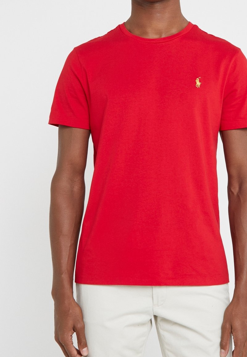 Rode katoenen T-shirt met een ronde hals, voorzien van een klein geel logo op de linkerborst. Gladde textuur, eenvoudig ontwerp.