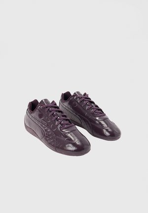 Paar dunkelviolette glänzende Sportschuhe mit Schnürsenkeln und dezenten kreisförmigen Mustern auf glatter Oberfläche vor hellem Hintergrund.