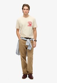 Beige bomulds T-shirt med et rødt grafisk design, parret med khaki corduroy bukser og en lyseblå stribet skjorte bundet om taljen. Brune sko.