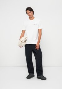 Witte katoenen T-shirt met een klein zwart logo, gecombineerd met donkere cargobroeken en zwarte sneakers, model dat een beige tas vasthoudt.