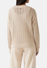 Vista posteriore di una persona che indossa un maglione a rete aperta di colore crema e pantaloni beige chiaro contro uno sfondo semplice.