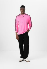 Long-sleeve pink athletic shirt with black three-stripe design on sleeves; paired with black pants and white sneakers. Simple, sporty style.
