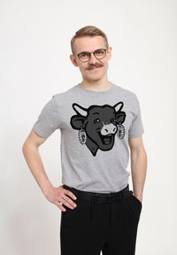 Graues Kurzarm-T-Shirt mit einem schwarzen Grafikdruck eines Kuhgesichts, das mit dekorativen Ohrringen geschmückt ist. Das Modell kombiniert es mit schwarzen Hosen.