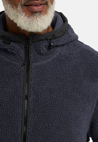 Giacca in pile di colore blu navy con superficie strutturata, dotata di chiusura con zip nera e cappuccio regolabile. Tessuto morbido e caldo con un design casual.
