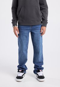 Sudadera con capucha de color gris oscuro con un bolsillo frontal, combinada con jeans de mezclilla azul claro y zapatillas negras con acentos y cordones blancos.