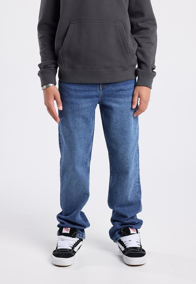 Sudadera con capucha de color gris oscuro con un bolsillo frontal, combinada con jeans de mezclilla azul claro y zapatillas negras con acentos y cordones blancos.