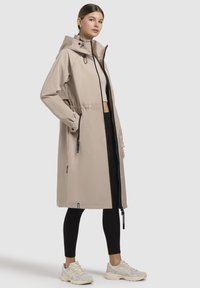 Palton trench waterproof bej cu glugă, talie elastică și fermoar în față, combinat cu colanți negri și adidași albi.