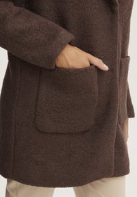 Manteau en mélange de laine marron avec un tissu texturé, présentant une poche avant et une coupe décontractée. Détail de couture visible près du col et de l'ourlet.