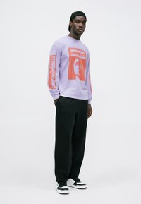 Langärmliges lila Shirt mit rotem Grafikdesign auf der Vorderseite und den Ärmeln, kombiniert mit lockeren schwarzen Hosen und schwarz-weißen Sneakers.