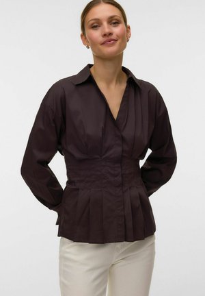Mujer con blusa plisada de color marrón oscuro con mangas largas y pantalones blancos, de pie frente a un fondo claro y liso.