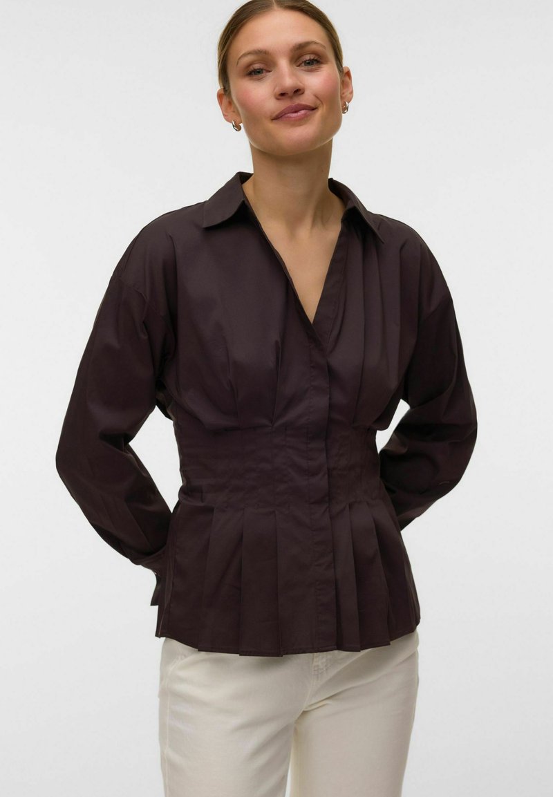 Femme portant un chemisier plissé marron foncé à manches longues et un pantalon blanc, debout devant un fond clair uni.
