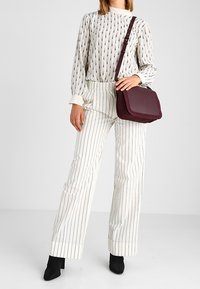 Borsa a spalla color mogano realizzata in materiale liscio, con un design strutturato e tracolla regolabile. Il modello indossa una blusa a fantasia e pantaloni a righe.